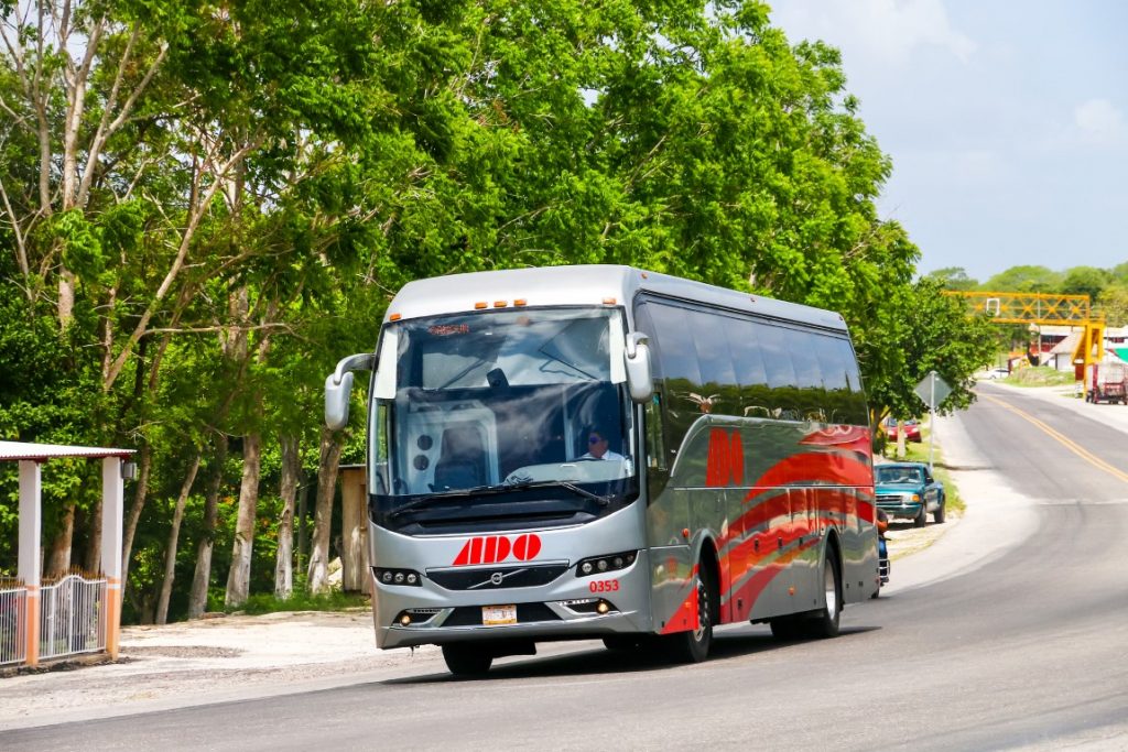 Image of a grey bus driving on a road with vividly green trees to the side. It's the ADO bus to get from Tulum to Holbox. 
