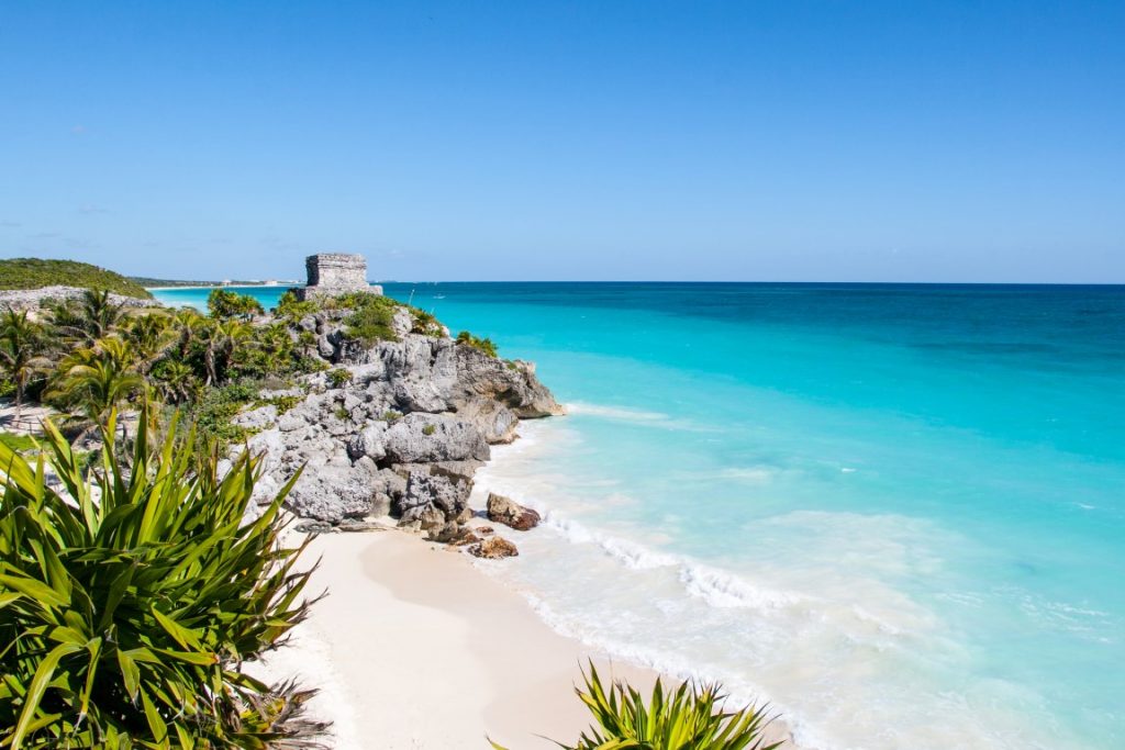 Image of Playa Ruinas, with the turquoise sea on the right, and a rocky cliffs with Mayan ruins on the left