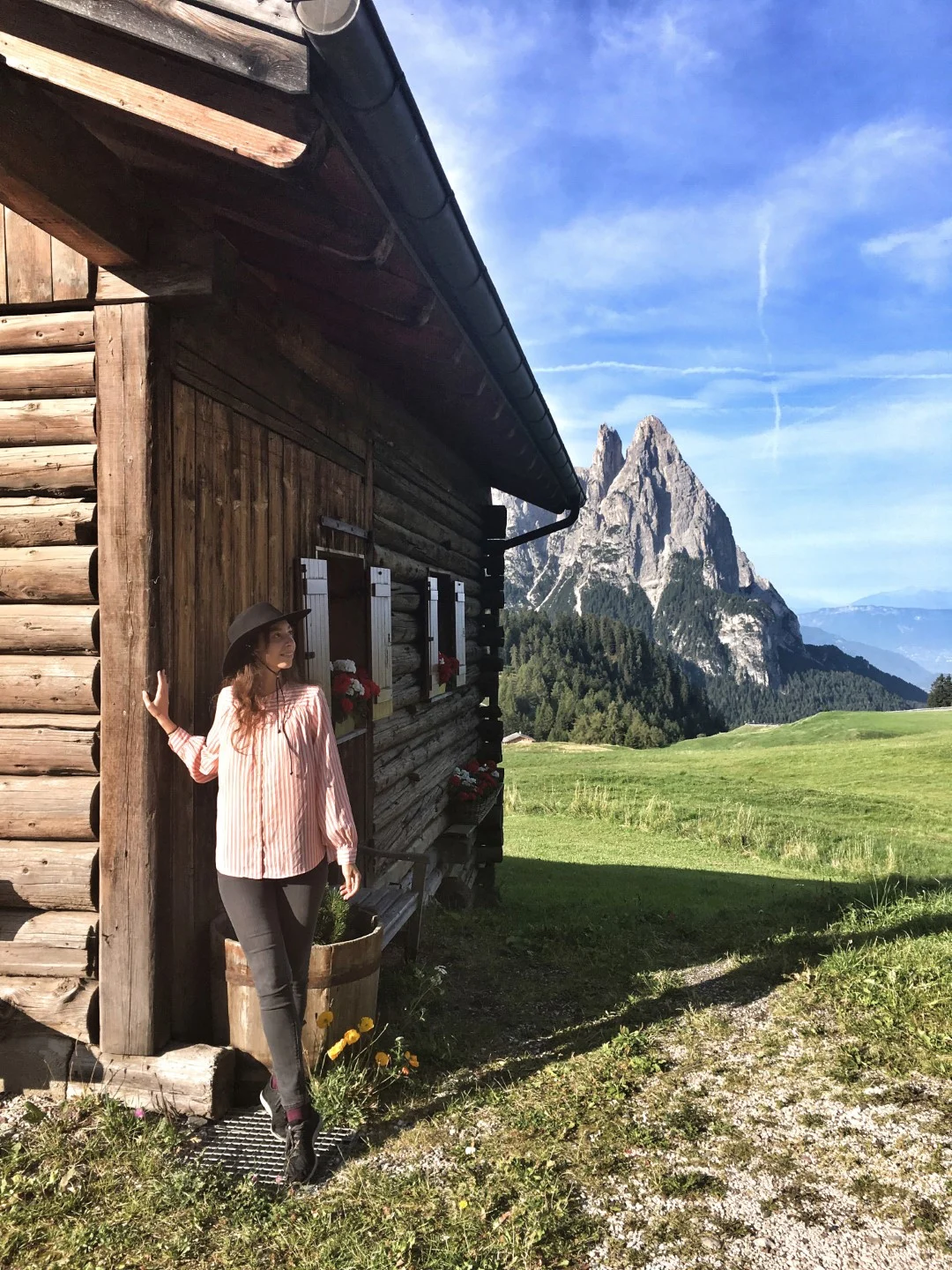 A woman posing outside a wooden cabin, with the striking Dolomite mountains in the background