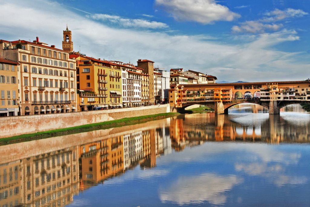 Image of a river with a colorful closed bridge above it, and vibrant buildings to the side, reflected on the water. It's the Arno river in Florence, a must on any Tuscany itinerary  