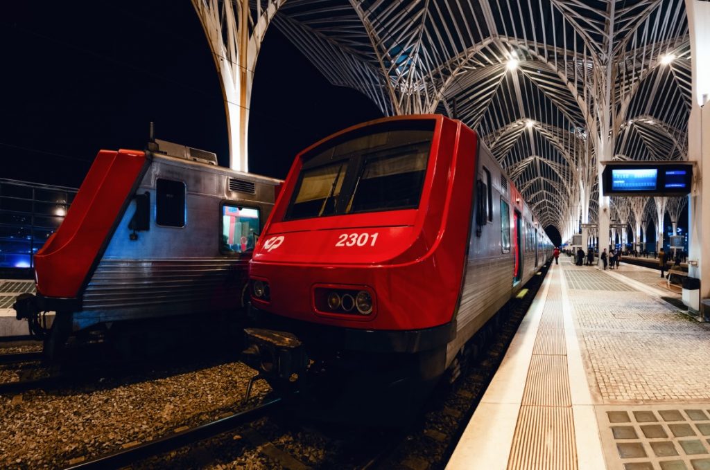 Image of a red train in a station in Italy, with a grey and red train behind it, and a platform with few people. Taking the train is the best way to make the day trip to Porto from Lisbon