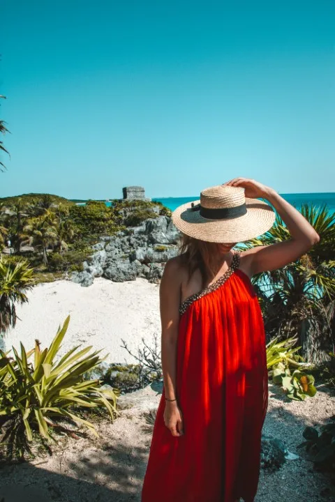 A woman wearing a red dress and a hat, standing at a viewpoint in the Mayan Ruins of Tulum, with a beach and a structure in the background