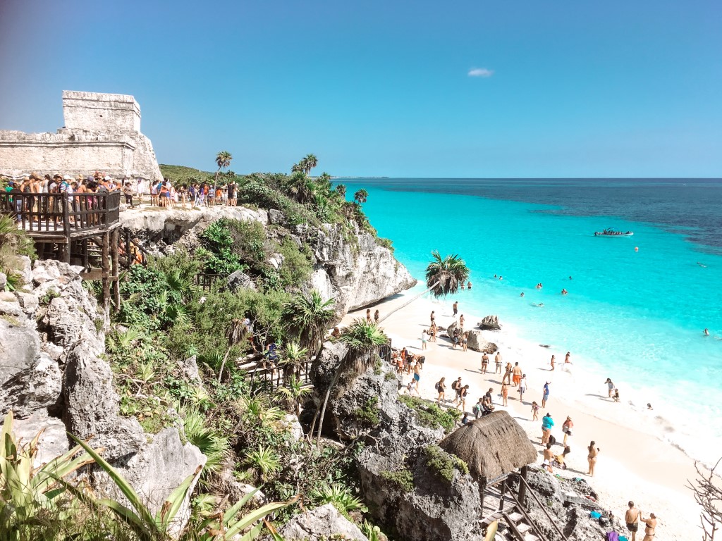 Image of Playa Ruinas, featuring a cliff with palm trees on the left, white sand with people standing, and turquoise waters at the bottom of Tulum Ruins, one of the unmissable stops on any Tulum itinerary
