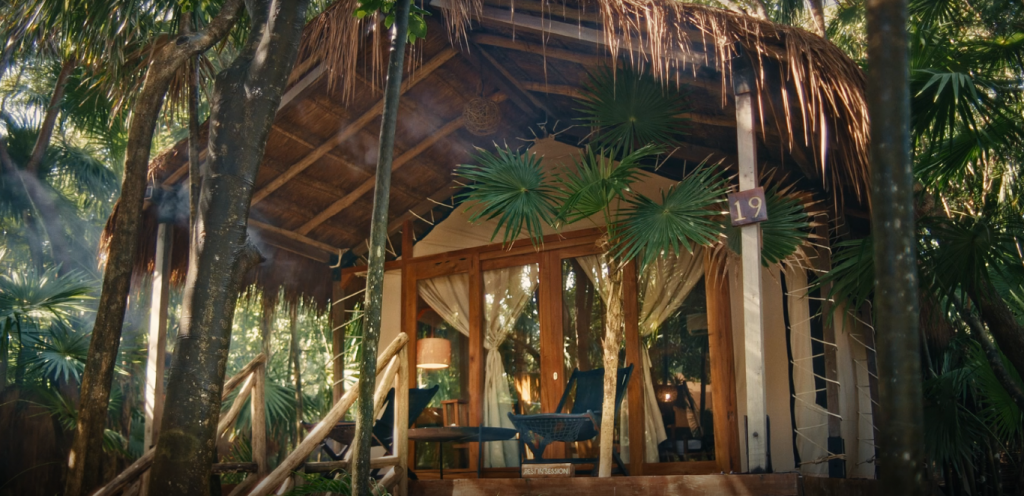 A thatched-roofed cabin with glass doors and a porch in the middle of the jungle, inserted in a post about Tulum eco hotels