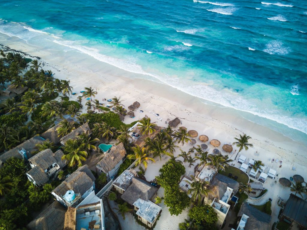 A drone shot featuring the turquoise waters of the Caribbean, a stretch of white sand, palm trees and palapas. Inserted in a post about taking a day trip to Tulum from Cancun
