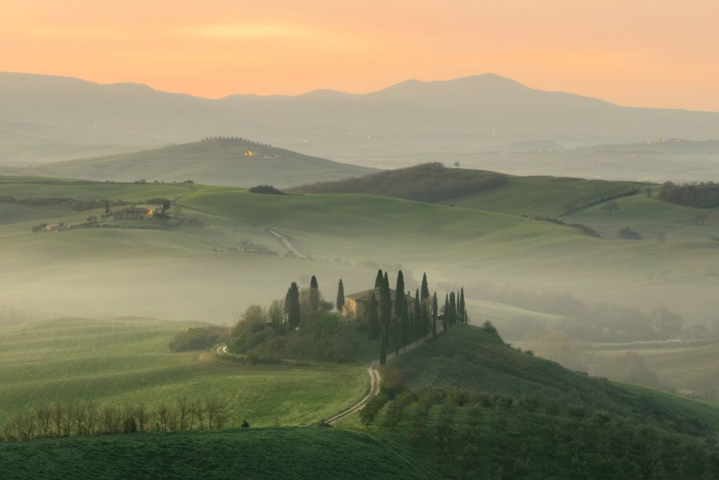 Image of Tuscany countryside from above, showing a road flanked by cypress trees, a villa, and rolling green fields around it, with mountains in the background, inserted in a post about a road trip Tuscany itinerary