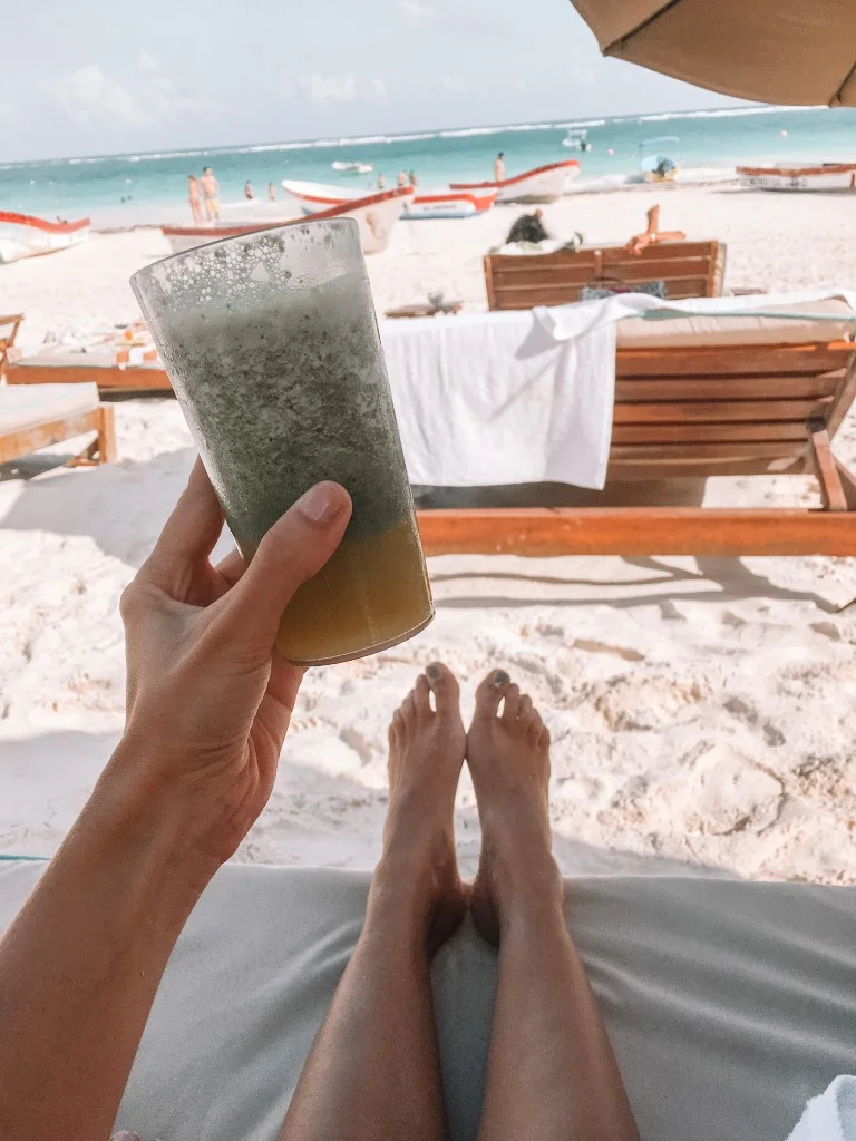 A girl's hand holding a smoothie at the beach, with sun chairs and boats in the background. Inserted in a post about taking a day trip to Tulum from Cancun
