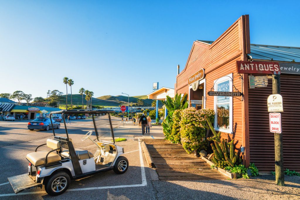 An image of an antiques shop, and a golf cart parked in front of it. In the background there are hills and palm trees. The image shows one of the best things to do in Cayucos: shopping for antiques