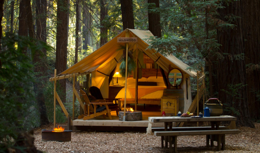 Image of a tent in a wooden deck, surrounded by forest. Inside the tent a bed, chairs, and a bedside nighttable can be seen. It's one of the best spots for glamping in Big Sur
