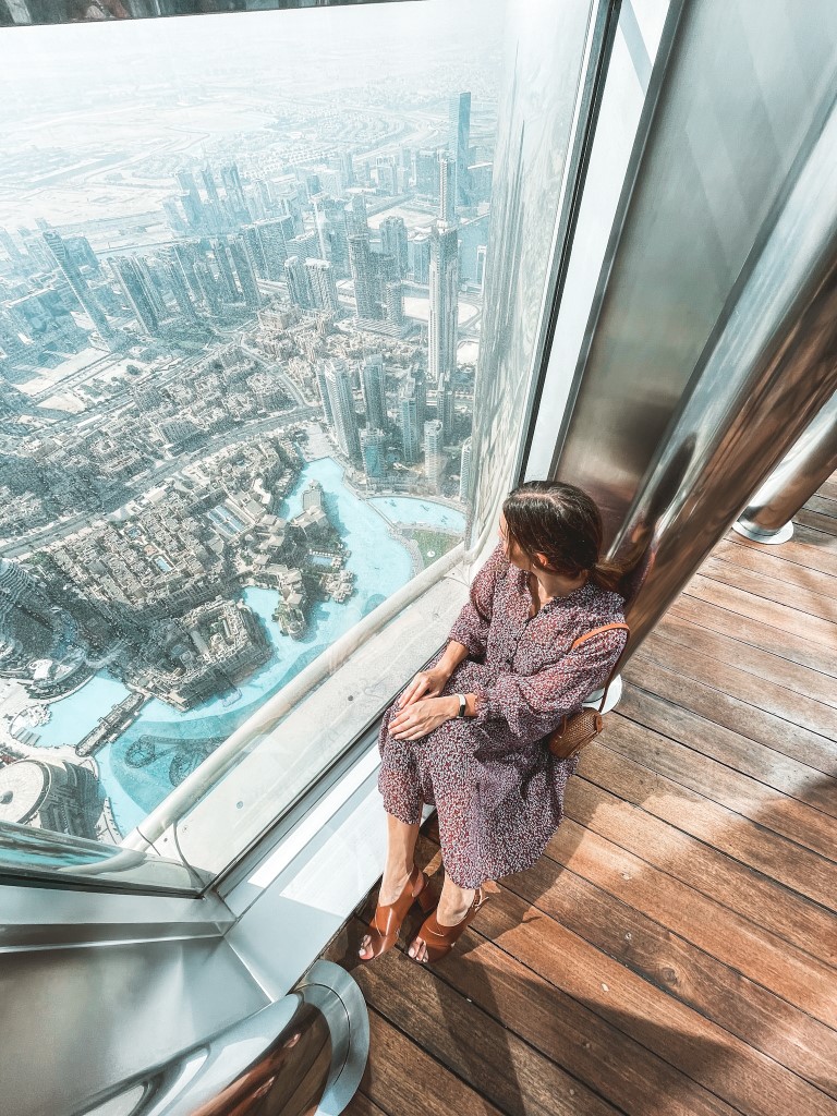 Image of a woman sitting by a window at Burj Khalifa, admiring the view of Dubai below. The picture is inserted in a post about Burj Khalifa facts