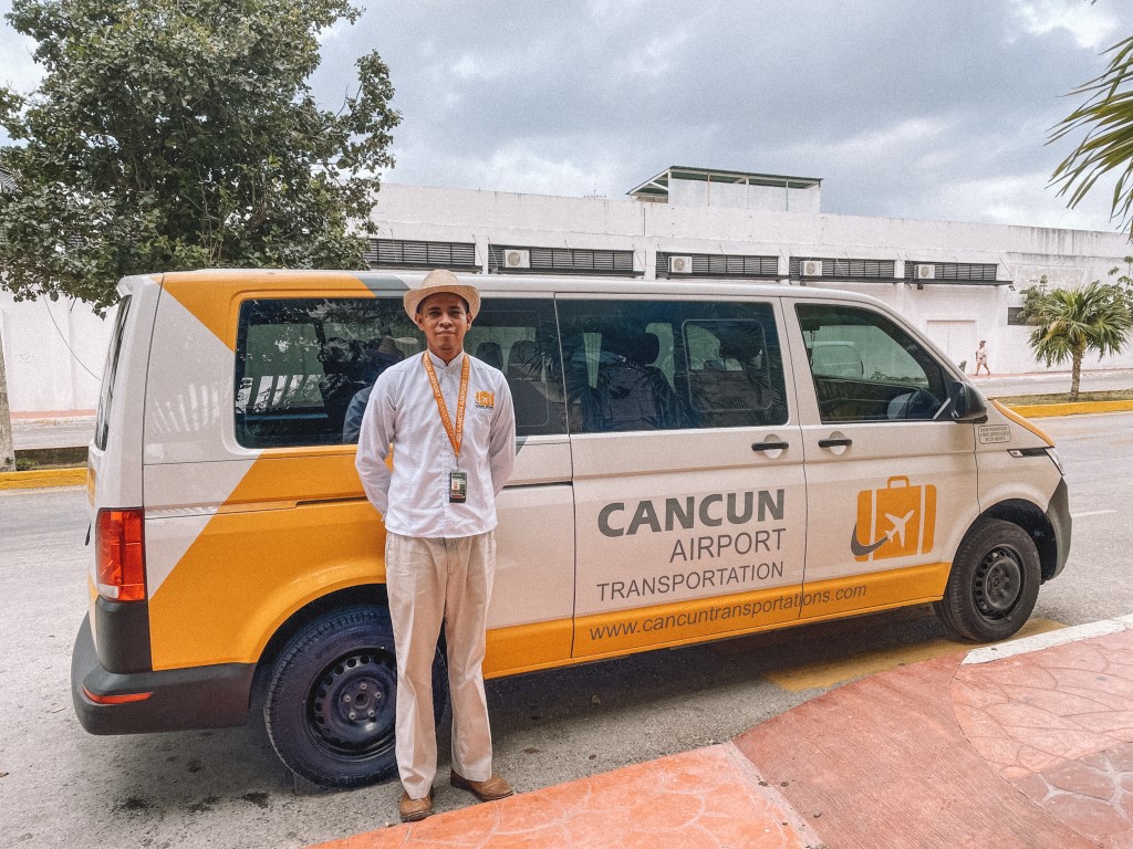 Image of a white and yellow van parked on the street, and the driver smiling in front of it. It's a shared shuttle, one of the best ways to get from Cancun Airport to Riviera Maya