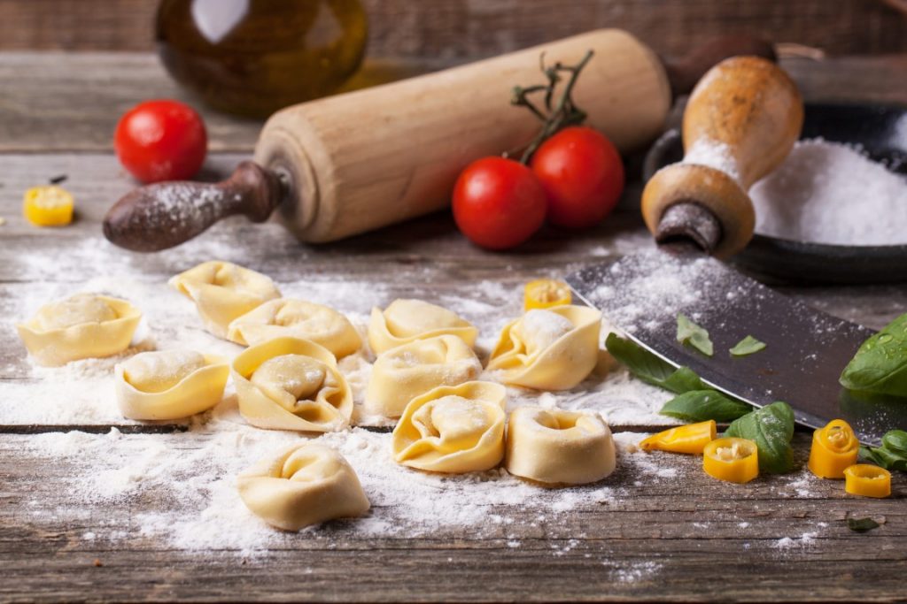 Fresh tortellini covered in flour on a wooden table. In the background there's a rolling pin, three red tomatoes, and a bowl of flour. Image inserted in a post about the best cooking classes in Bologna