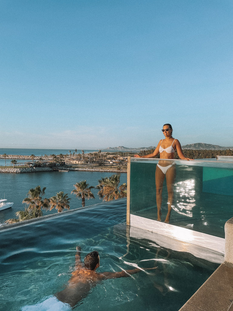 A woman in a white bikini standing in a glass pool, while a man swims in the swimming pool right below, and views of Los Cabos in the background