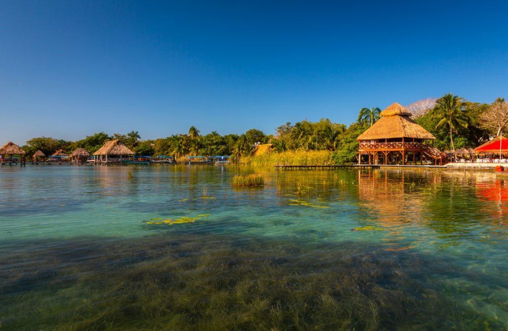 Image of Bacalar lagoon, with palm trees and thatched-roof cabins on the shore, inserted in a post about getting from Tulum to Bacalar