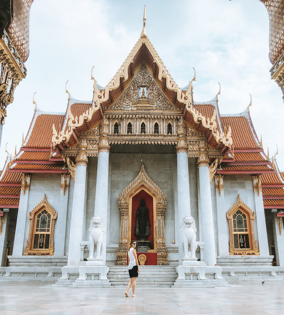 Image of a white temple with golden windows and door, and a woman standing in front of it inserted in a post about the best temples in Bangkok