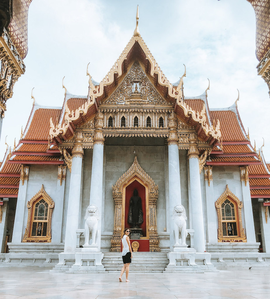 Image of a white temple with golden windows and door, and a woman standing in front of it inserted in a post about the best temples in Bangkok 