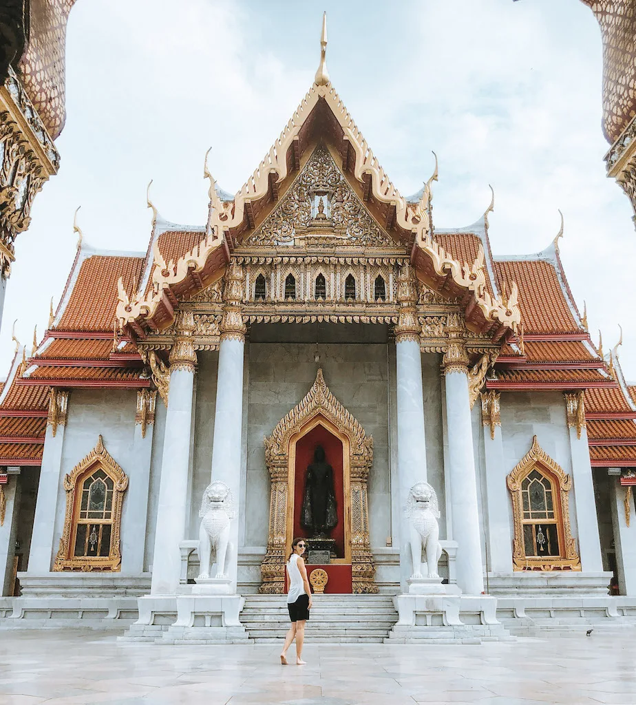Image of a white temple with golden windows and door, and a woman standing in front of it inserted in a post about the best temples in Bangkok 