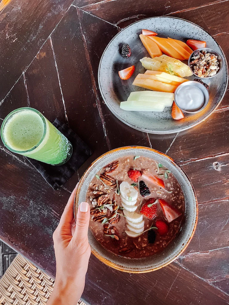 A bowl of oatmeal with fruit on top, a plate of fruit, and a green smoothie on a breakfast table in Hotel El Ganzo