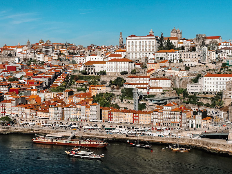 White and light-colored buildings with terracota roofs scattered in the image, with the river at the bottom and 4 ships by the boardwalk next to the river, inserted in a post about the perfect day trip to Porto from Lisbon 