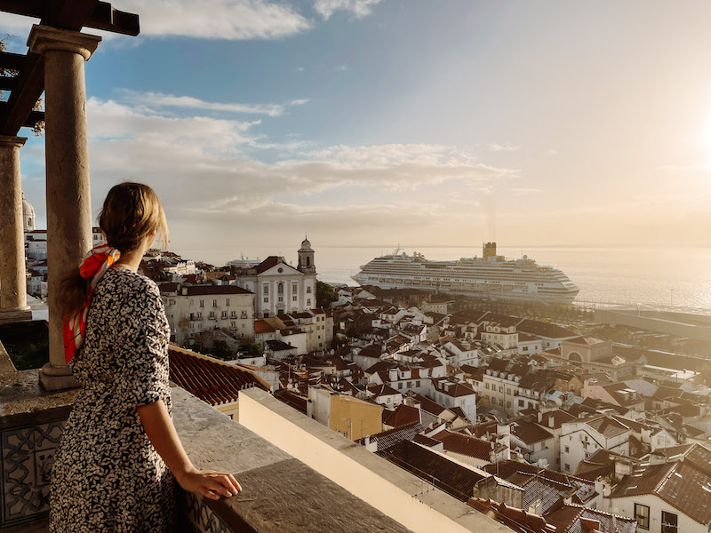 A girl looking at the terracota rooftops of Lisbon from a viewpoint, with the river and a cruise in the background. Image inserted in a post about the best 3 days in Lisbon itinerary