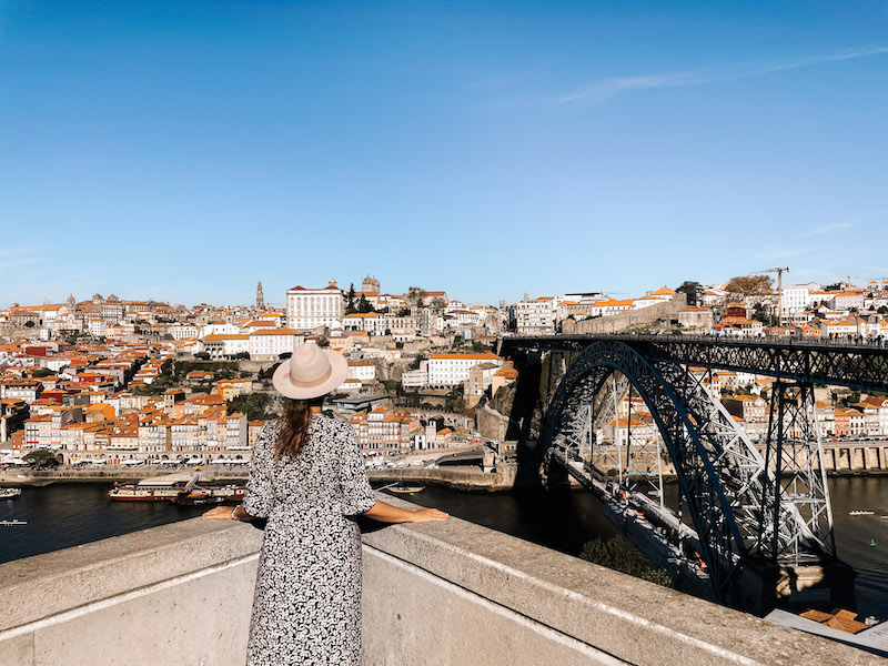 Image of a woman looking at the white buildings with terracotta rooftops across the river, with Dom Luis Bridge to the right, a great activity to enjoy while spending one day in Lisbon