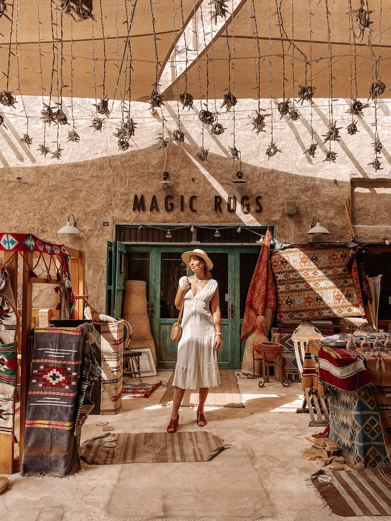 A woman in a white dress standing in front of a carpet shop, surrounded by carpets of different colors and designs, in one of the souks in Dubai