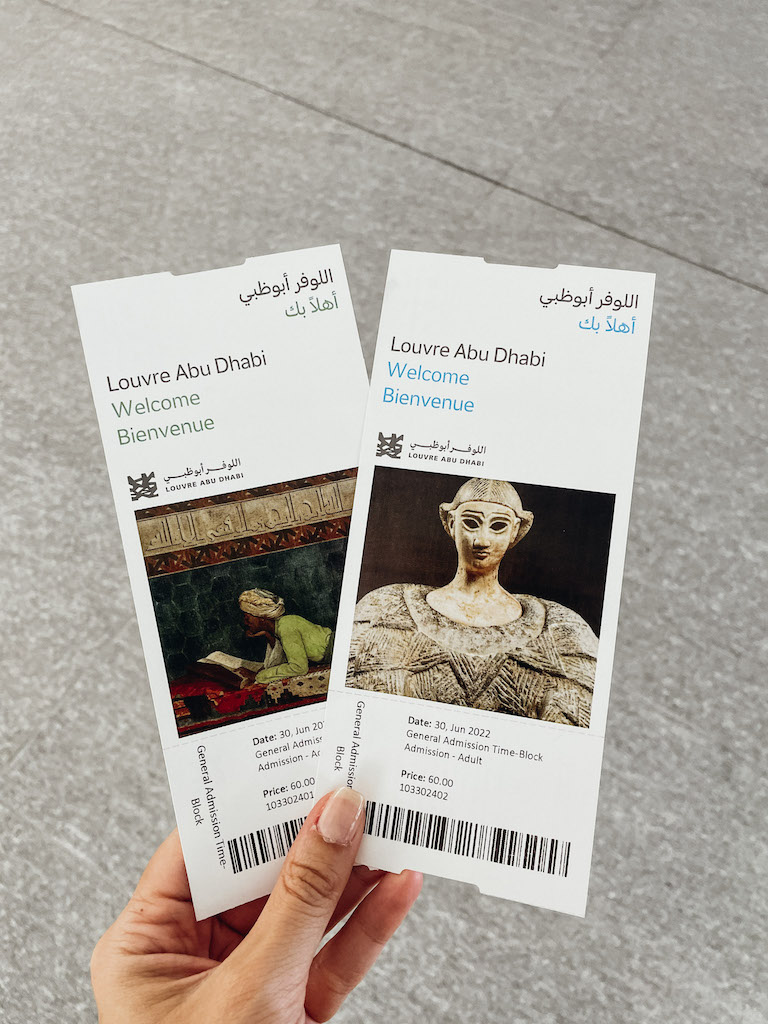 Two The Louvre Abu Dhabi Tickets being hold by a woman's hand, against the grey floor of the museum. 