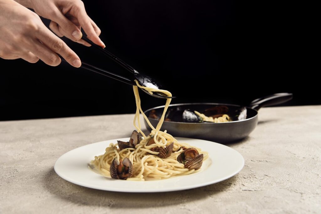 Image of spaghetti with seafood in a white rounded plate, with a black pan in the background, inserted in a post about the best cooking classes in Venice