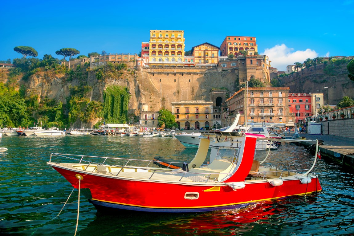 Image of a red boat on the water, with an impressive cliff and colorful houses in the background