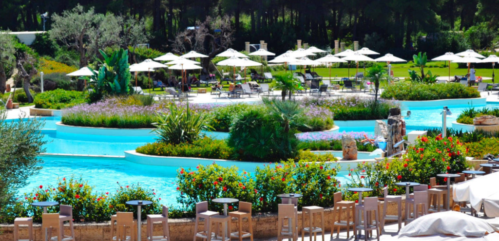 Image of the swimming pool area of Vivosa Apulia Resort, surrounded by plants and with white umbrellas in the background