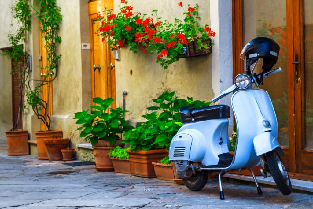 Image of a small street with flowers and plants against yellow walls, a rustic entrance, and a blue Vespa standing in front of the door, inserted in a post about Florence Vespa Tours 