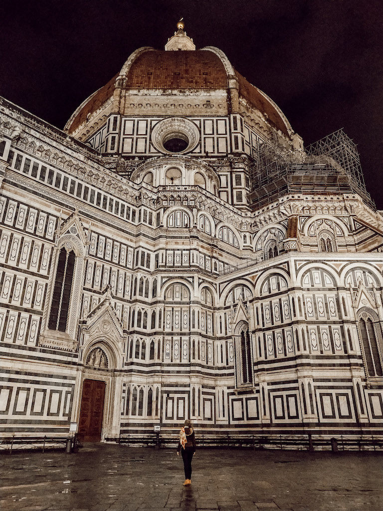 Image of the Cathedral of Santa Maria del Fiore and the impressive Brunelleschi's Duomo on top at night, with a woman admiring it from the square.