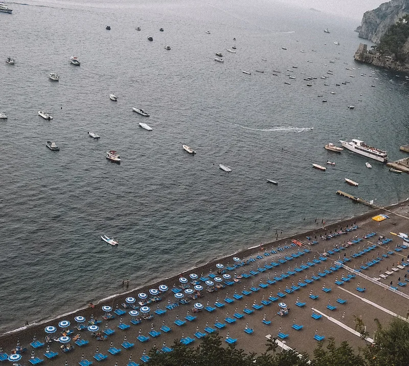 Image of a beach in Positano seen from above, with blue sun loungers and umbrellas on the shore and boats dotting the sea.