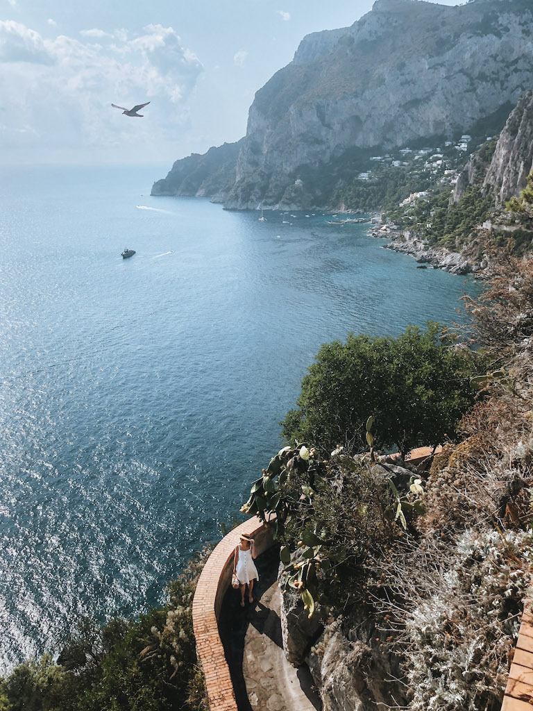 A woman posing in the very narrow Vis Tragari, and in the background there's the Mediterranean Sea, and the rugged coastline of Capri island 