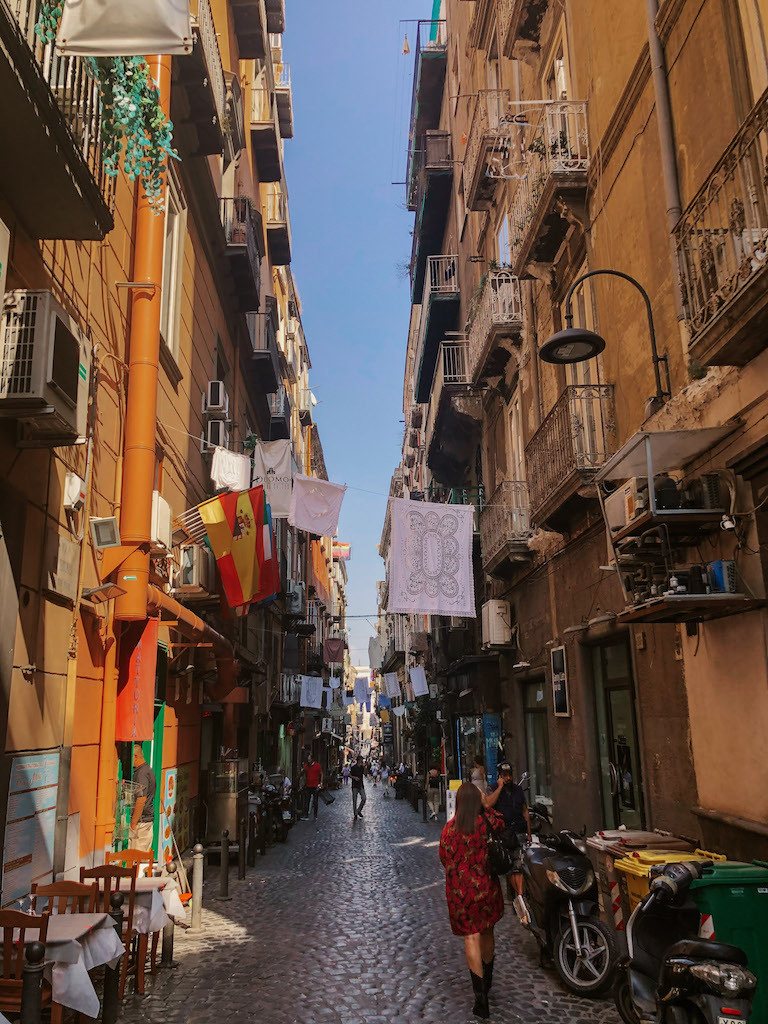 A narrow cobblestone street sandwiched between two buildings. The street is lined by restaurant tables and motorbikes, and there's laundry hanging between the balconies on both sides. 