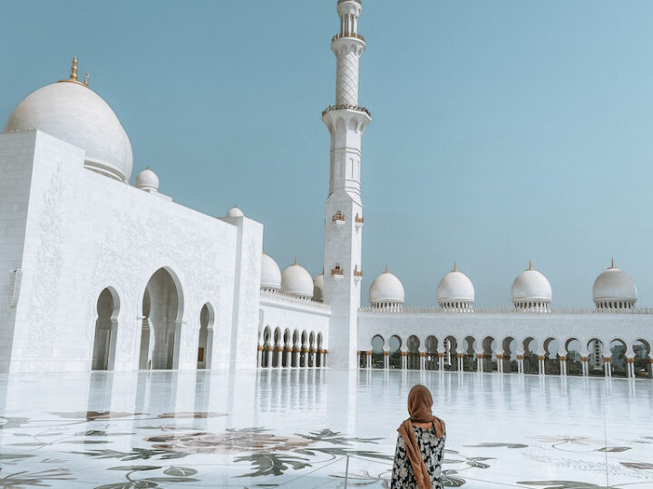 Woman from the back wearing an Arabic-style dress and a scarf on her head is standing in the courtyard and looking at the majestic white Grand Zayed Mosque during a day tour from Dubai to Abu Dhabi.