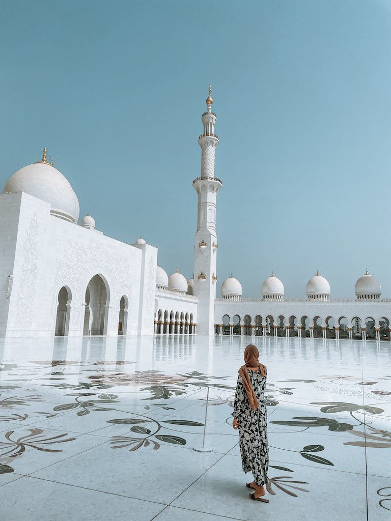 Woman from the back wearing an Arabic-style dress and a scarf on her head is standing in the courtyard and looking at the majestic white Grand Zayed Mosque during a day tour from Dubai to Abu Dhabi. 