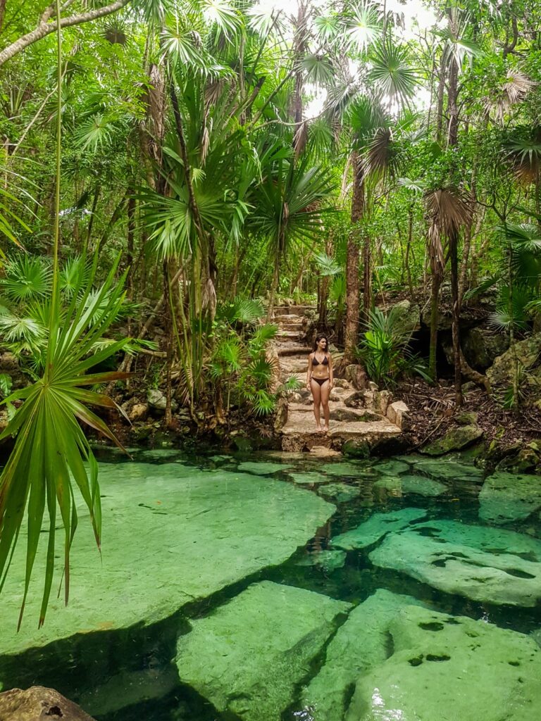 A photograph of a woman standing at the end of a stone pathway leading to Cenote Azul, taken from the opposite side of the cenote