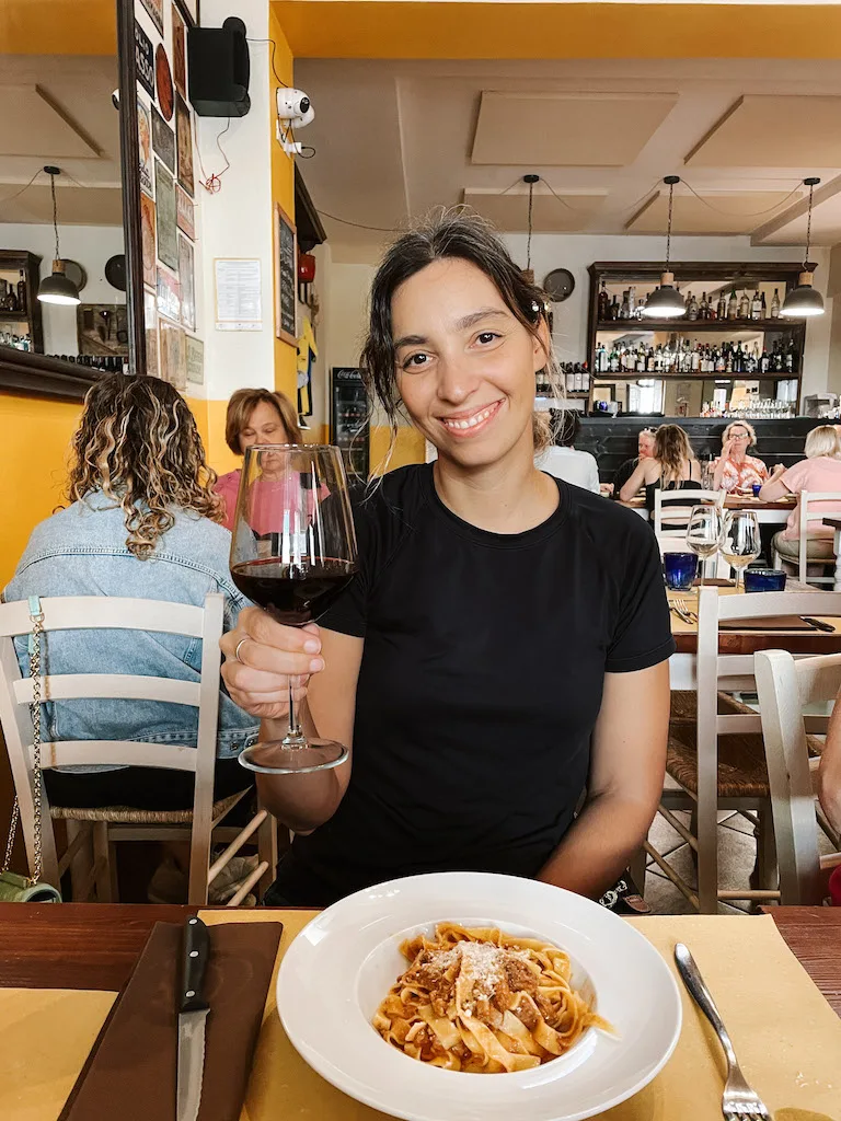 A woman smiling to camera, holding a wine glass on one hand. There's a plate of tagliatelle al ragu in front of her