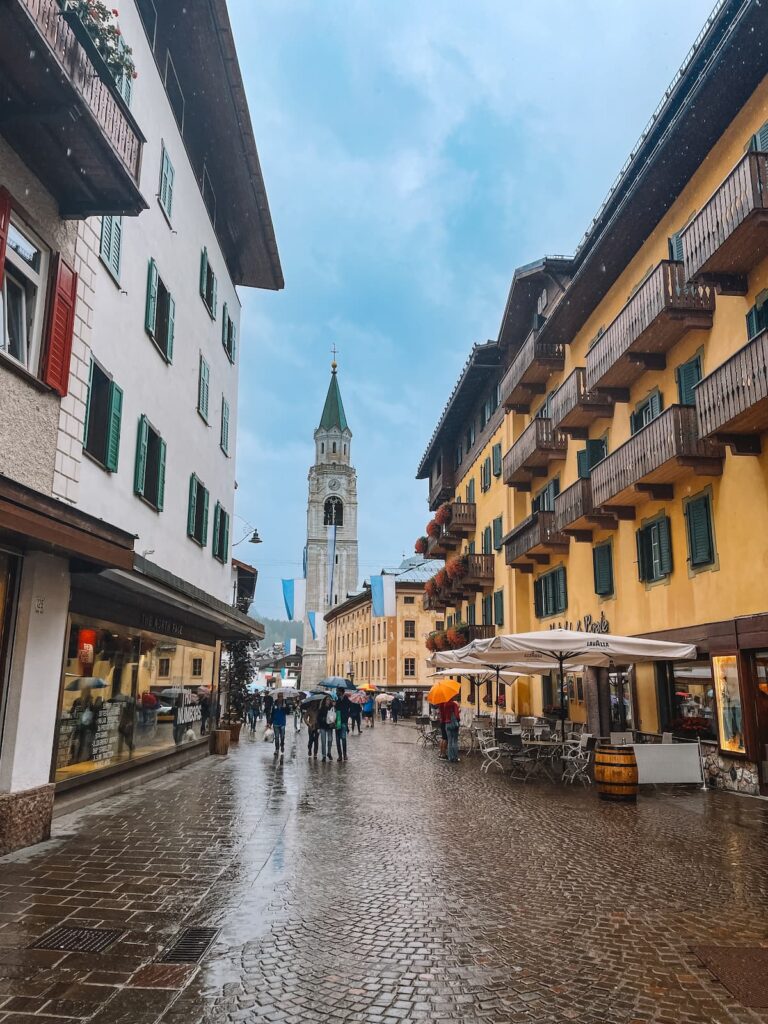 Rainy street in Cortina d'Ampezzo, with colorful alpine buildings, café tables, umbrellas, and the church bell tower in the background.