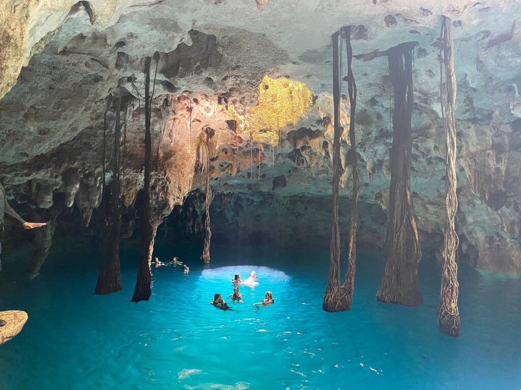 The inside of a cave cenote with stalactites, a few people swimming in the blue water.