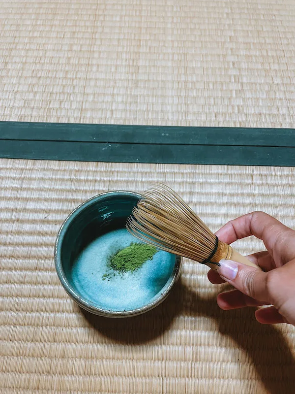 A matcha bowl with tea powder inside, and a woman's hand holding a matcha whisk