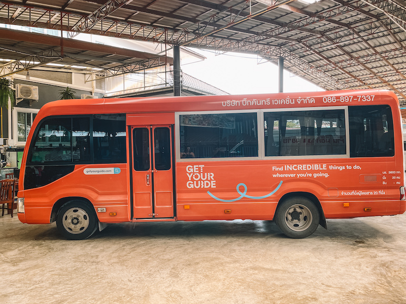 "Orange shuttle bus with 'Get Your Guide' branding parked under a covered area in Bangkok.