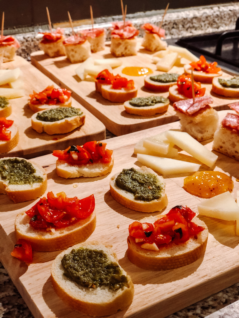 Wooden boards filled with appetizers like bruschette with pesto and tomatoes, cheese slices, salami on bread, and jam.