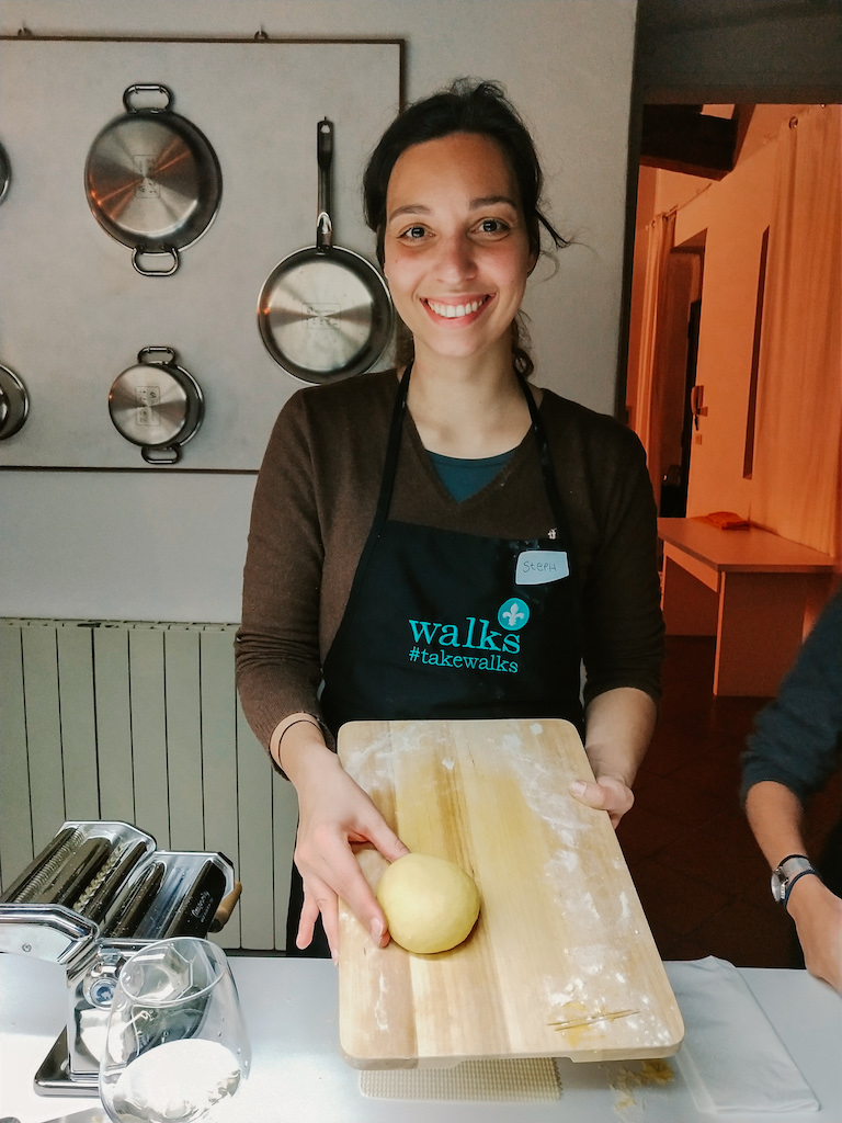 Smiling woman wearing a black apron and holding a cutting board with fresh pasta dough in a kitchen