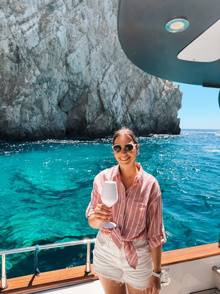Woman smiling on a boat tour in Capri with a drink in hand and turquoise water by the cliffs behind her