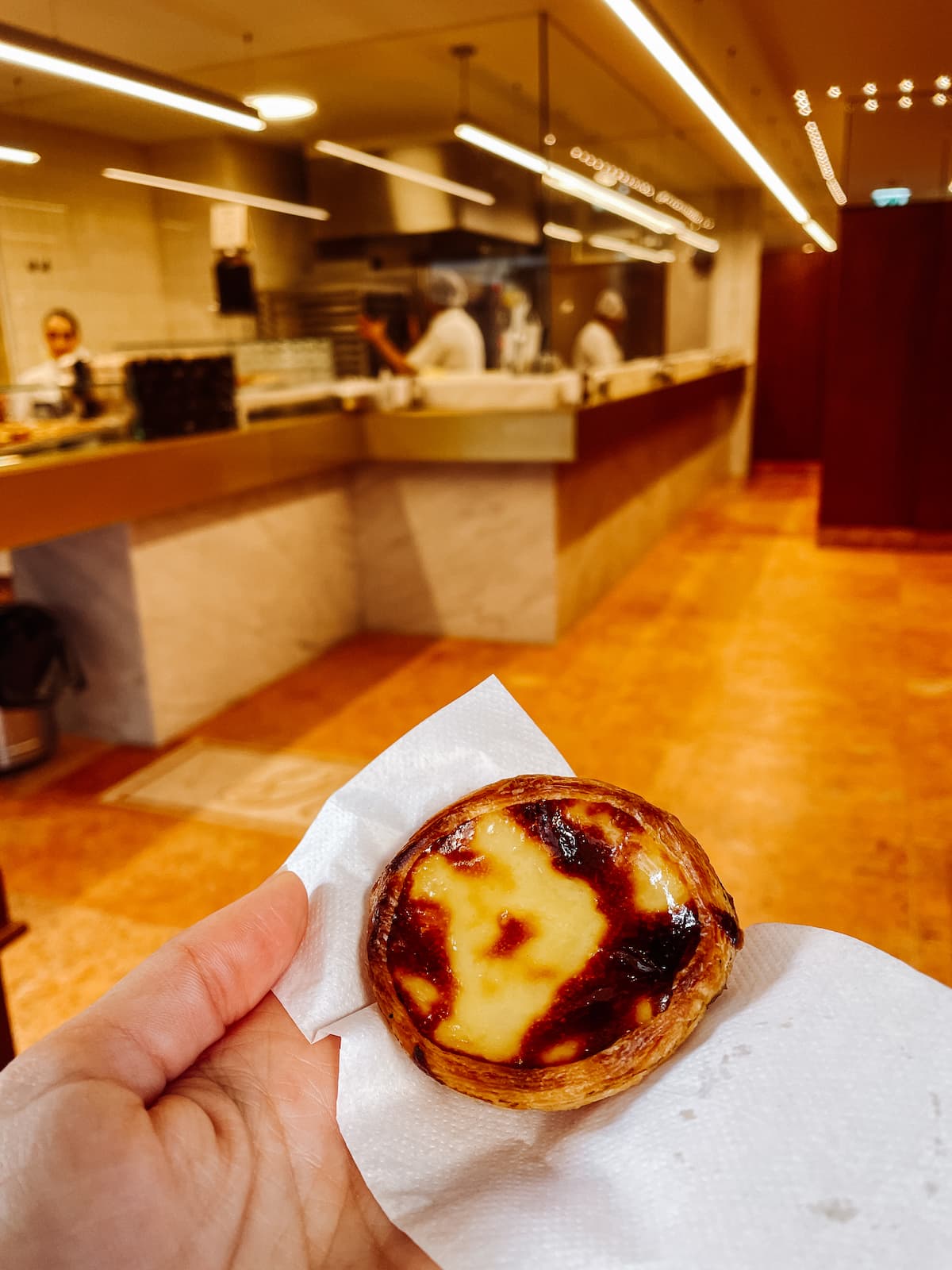 Hand holding a freshly baked pastel de nata in a Porto bakery with an open kitchen in the background