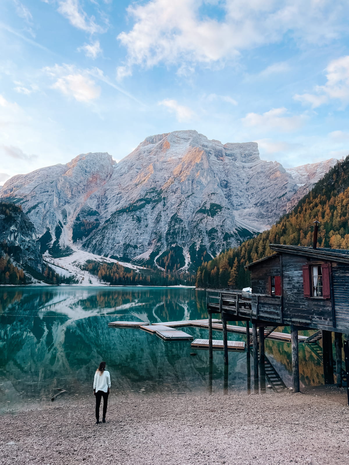 Woman standing on the shore of Lake Braies, with turquoise water, a wooden boathouse, and dramatic Dolomites peaks in the background.