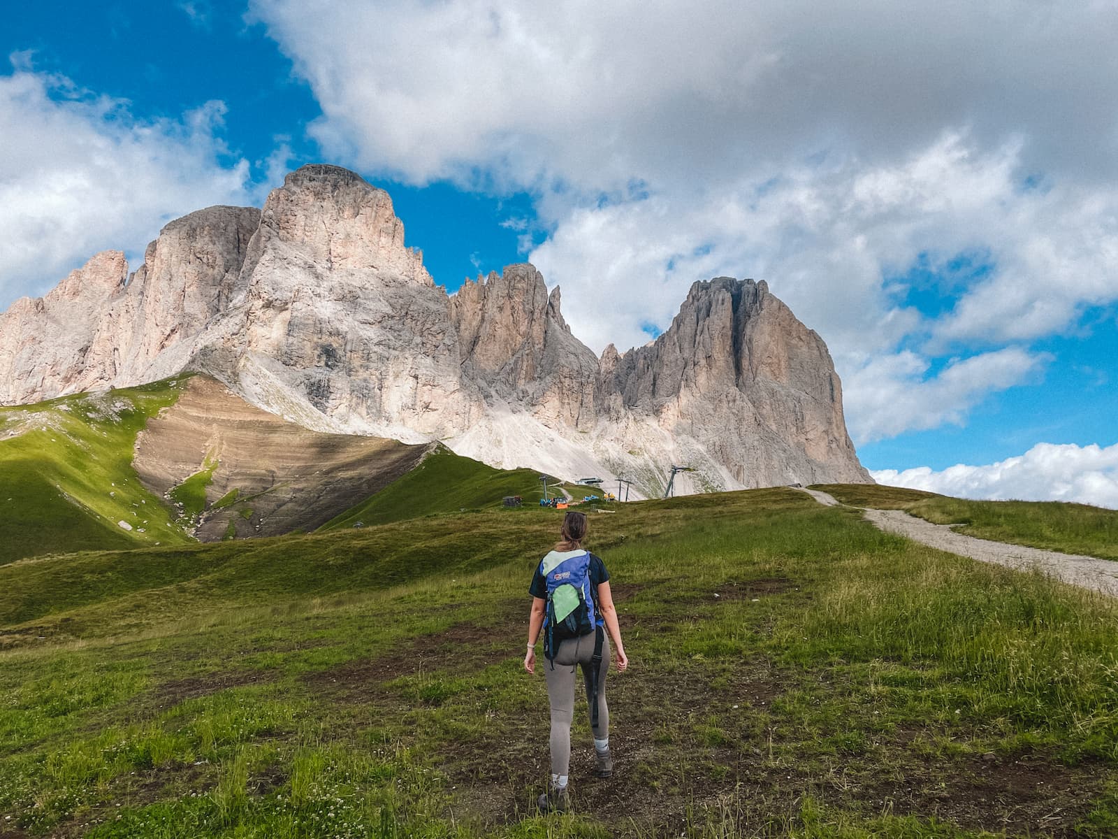 Woman hiking on a grassy trail in the Dolomites, with jagged mountain peaks and a cloudy blue sky ahead.