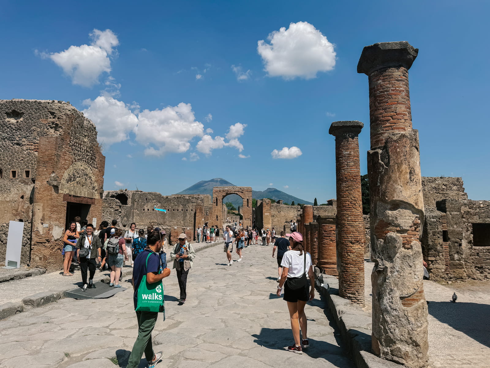 Ruins of Pompeii with visitors walking along the ancient stone street, brick columns in the foreground, and Mount Vesuvius in the background.